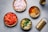 Several bowls are arranged on a surface, each filled with different cooking ingredients. There are chopped onions, diced tomatoes, an assortment of ground spices, chopped green chilies with garlic, and a bottle of oil. A grater is positioned nearby, completing the cooking setup.