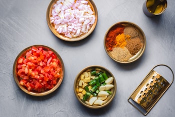 Several bowls are arranged on a surface, each filled with different cooking ingredients. There are chopped onions, diced tomatoes, an assortment of ground spices, chopped green chilies with garlic, and a bottle of oil. A grater is positioned nearby, completing the cooking setup.