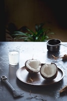 A wooden plate holds two halves of a coconut, creating a rustic and natural setting. Nearby, a glass filled with coconut water stands next to a metal tool, possibly for cracking coconuts. A small pot is also present on the table, and a potted plant in the background adds a touch of greenery.