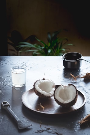 A wooden plate holds two halves of a coconut, creating a rustic and natural setting. Nearby, a glass filled with coconut water stands next to a metal tool, possibly for cracking coconuts. A small pot is also present on the table, and a potted plant in the background adds a touch of greenery.