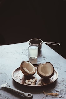A coconut split into halves is placed on a round tray, with small coconut pieces arranged around it. In the background, a glass filled with coconut water and a strainer are visible. The surface is slightly wet, with an adjustable wrench lying nearby, among coconut husk strands.