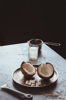A coconut split into halves is placed on a round tray, with small coconut pieces arranged around it. In the background, a glass filled with coconut water and a strainer are visible. The surface is slightly wet, with an adjustable wrench lying nearby, among coconut husk strands.