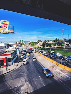 A busy urban street scene with a large traffic jam. Cars are lined up on the road, moving slowly. A billboard advertising a corn product, 'Famosa Maíz con Coco,' is prominently displayed on the left side. Buildings and commercial establishments flank the street. A few pedestrians are walking on the sidewalk. Electrical wires crisscross above, and a clear blue sky forms the backdrop.