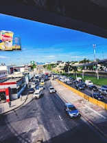 A busy urban street scene with a large traffic jam. Cars are lined up on the road, moving slowly. A billboard advertising a corn product, 'Famosa Ma&iacute;z con Coco,' is prominently displayed on the left side. Buildings and commercial establishments flank the street. A few pedestrians are walking on the sidewalk. Electrical wires crisscross above, and a clear blue sky forms the backdrop.