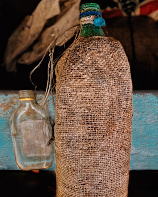 A large green bottle wrapped in a coarse brown burlap material stands upright next to a smaller, clear glass bottle. Blue and white twine is tied around the neck of the green bottle, and both bottles are set against a textured blue wooden surface with a dark, blurred background.
