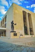 A statue of a person holding a book stands in front of a large, beige stone building with tall, arched windows. The architecture is modern with straight lines and a clean design. The sky above is a vibrant blue with scattered white clouds.
