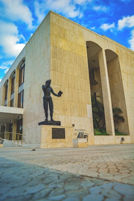 A statue of a person holding a book stands in front of a large, beige stone building with tall, arched windows. The architecture is modern with straight lines and a clean design. The sky above is a vibrant blue with scattered white clouds.