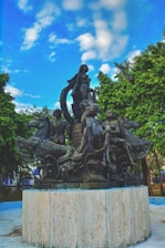 A detailed sculpture featuring multiple human figures and mythical creatures situated on a circular stone pedestal. The artwork is surrounded by lush green trees under a vibrant sky with scattered clouds.