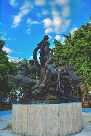 A detailed sculpture featuring multiple human figures and mythical creatures situated on a circular stone pedestal. The artwork is surrounded by lush green trees under a vibrant sky with scattered clouds.