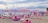 A beach scene with numerous people relaxing on variously colored beanbags under pastel-toned umbrellas. The beach is lined with string lights, and there are inflatable water structures in the sea. The sky is overcast with clouds.