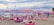 A beach scene with numerous people relaxing on variously colored beanbags under pastel-toned umbrellas. The beach is lined with string lights, and there are inflatable water structures in the sea. The sky is overcast with clouds.