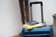 A blue suitcase with a retractable handle is positioned in a well-lit area. On top of the suitcase, there is a hardcover book with a yellow cover and a closed passport resting on it.