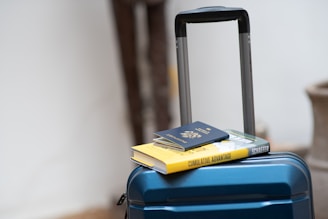 A blue suitcase with a retractable handle is positioned in a well-lit area. On top of the suitcase, there is a hardcover book with a yellow cover and a closed passport resting on it.