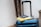 A blue suitcase with a retractable handle is positioned in a well-lit area. On top of the suitcase, there is a hardcover book with a yellow cover and a closed passport resting on it.