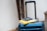 A blue suitcase with a retractable handle is positioned in a well-lit area. On top of the suitcase, there is a hardcover book with a yellow cover and a closed passport resting on it.