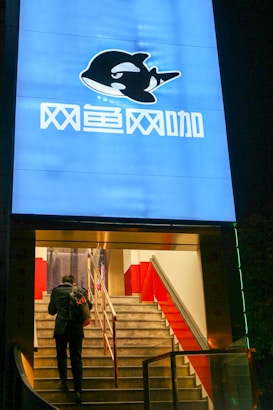 The image features a brightly lit entrance to a cybercafe with a large blue sign displaying an orca logo and Chinese characters. A person carrying a backpack is ascending a staircase leading to the entrance, with red railings on each side.