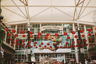 An entrance to a shopping complex adorned with numerous red lanterns and a decorative dragon suspended in the air. The structure is a large, modern building with glass windows and a fabric canopy overhead. A sign reads 'Hua Hin Market Village' in both English and Thai. The atmosphere is festive, suggesting a celebration or cultural event.