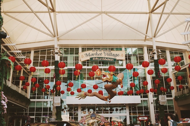 An entrance to a shopping complex adorned with numerous red lanterns and a decorative dragon suspended in the air. The structure is a large, modern building with glass windows and a fabric canopy overhead. A sign reads 'Hua Hin Market Village' in both English and Thai. The atmosphere is festive, suggesting a celebration or cultural event.