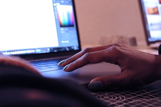 A person's hand is resting on the touchpad of a laptop, suggesting interaction with the device. The screen displays a bright interface, likely a graphic design or editing program. In the background, there is another computer screen visible, partially blurred.
