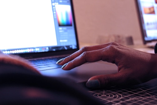 A person's hand is resting on the touchpad of a laptop, suggesting interaction with the device. The screen displays a bright interface, likely a graphic design or editing program. In the background, there is another computer screen visible, partially blurred.