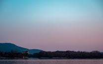 A serene landscape featuring a large body of water reflecting the soft pastel colors of a sunset or sunrise. In the distance, an island with lush trees and a small building on stilts is visible. The horizon is adorned with hills that gradually fade into the blue and pink sky.