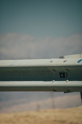 A metal guardrail is positioned horizontally across the image, with visible bolts and a slightly rustic texture. The background features a blurred landscape of hills and a blue sky, creating a muted, serene backdrop.