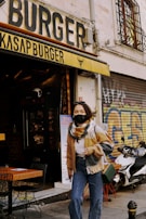 A person wearing a mask, scarf, and casual clothing stands in front of a burger shop named 'Kasap Burger' with bold signage. The shop has wooden outdoor tables and chairs, and a menu board is displayed at the entrance. A scooter is parked nearby, and the area features some street art graffiti on the walls.