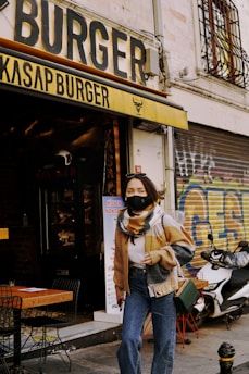 A person wearing a mask, scarf, and casual clothing stands in front of a burger shop named 'Kasap Burger' with bold signage. The shop has wooden outdoor tables and chairs, and a menu board is displayed at the entrance. A scooter is parked nearby, and the area features some street art graffiti on the walls.