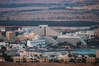 A sprawling cityscape featuring a dense cluster of large, modern buildings, including hotels and residential structures, set against a backdrop of hills and sparse vegetation. The architecture is dominated by light-colored facades, with distinctive geometric patterns. A large body of water is visible to the right, suggesting a coastal or waterfront location.