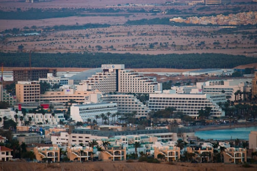 A sprawling cityscape featuring a dense cluster of large, modern buildings, including hotels and residential structures, set against a backdrop of hills and sparse vegetation. The architecture is dominated by light-colored facades, with distinctive geometric patterns. A large body of water is visible to the right, suggesting a coastal or waterfront location.