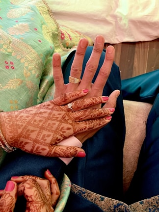 Two hands with intricate mehndi designs are placed together, displaying rings on the fingers. The background includes a richly decorated cloth with floral patterns. The nails are painted pink, and the person is wearing a traditional garment.