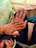 Two hands with intricate mehndi designs are placed together, displaying rings on the fingers. The background includes a richly decorated cloth with floral patterns. The nails are painted pink, and the person is wearing a traditional garment.
