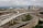 Aerial view of a complex highway interchange with multiple overpasses and underpasses, surrounded by urban buildings and skyscrapers in the background. The roads are under construction with visible concrete work and equipment scattered throughout the site. The sky is overcast, casting a dull light over the area.