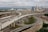 Aerial view of a complex highway interchange with multiple overpasses and underpasses, surrounded by urban buildings and skyscrapers in the background. The roads are under construction with visible concrete work and equipment scattered throughout the site. The sky is overcast, casting a dull light over the area.