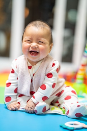 A joyful baby wearing a white outfit with large red polka dots sits on a colorful playmat, smiling brightly. The background is softly blurred, highlighting the cheerful expression.
