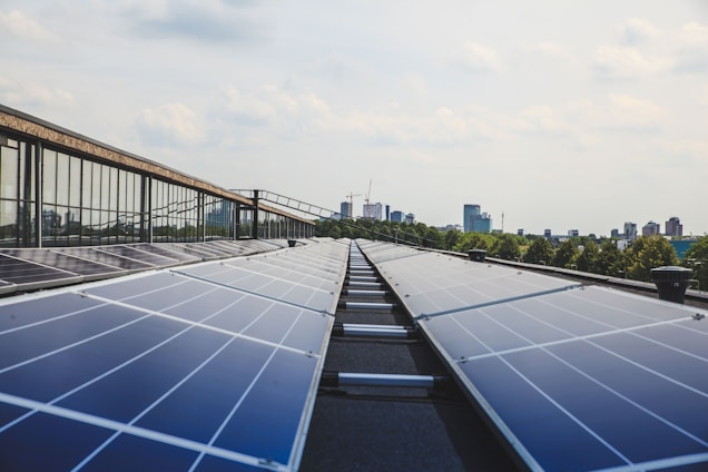 A row of solar panels on a rooftop stretches out towards the horizon, with reflections visible on their surfaces. In the background, a city skyline with high-rise buildings is partially obscured by trees. The sky is partly cloudy with soft, diffuse lighting.