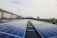A row of solar panels on a rooftop stretches out towards the horizon, with reflections visible on their surfaces. In the background, a city skyline with high-rise buildings is partially obscured by trees. The sky is partly cloudy with soft, diffuse lighting.