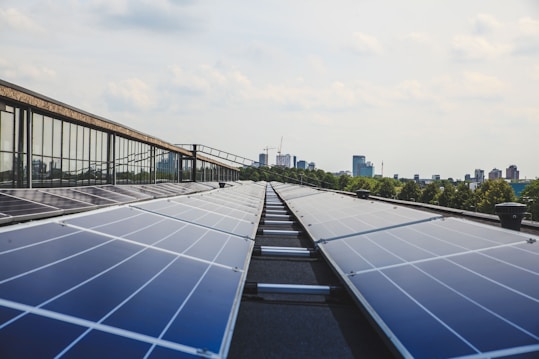 A row of solar panels on a rooftop stretches out towards the horizon, with reflections visible on their surfaces. In the background, a city skyline with high-rise buildings is partially obscured by trees. The sky is partly cloudy with soft, diffuse lighting.