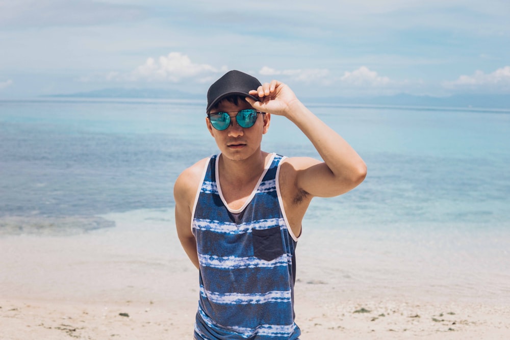 man in blue and white tank top wearing black sunglasses standing on beach during daytime