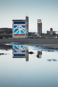 A large industrial building with a prominent mural painted on the side, depicting geometric shapes and lines in blue, red, and white. The structure is reflected in a still body of water in the foreground, enhancing the symmetry of the scene. Other industrial structures and a hilly landscape are visible in the background.