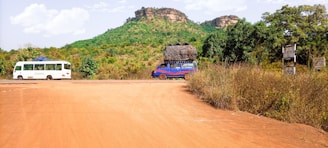 A rural scene with a dirt road in the foreground, flanked by lush green vegetation. In the background, a small rocky hill rises with sparse greenery on its surface. Two vehicles travel on the road: a white bus on the left and a blue van carrying a large load on its roof on the right. The sky is partly cloudy, indicating a clear day.