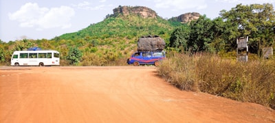 A rural scene with a dirt road in the foreground, flanked by lush green vegetation. In the background, a small rocky hill rises with sparse greenery on its surface. Two vehicles travel on the road: a white bus on the left and a blue van carrying a large load on its roof on the right. The sky is partly cloudy, indicating a clear day.