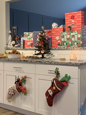 A countertop displays a collection of wrapped gifts in colorful holiday-themed paper. A small decorated Christmas tree is visible, adorned with ornaments and a star on top. Along the cabinets below are festive Christmas stockings hung with care. A variety of baked goods are arranged at one end of the counter, suggesting a holiday gathering.