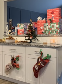 A countertop displays a collection of wrapped gifts in colorful holiday-themed paper. A small decorated Christmas tree is visible, adorned with ornaments and a star on top. Along the cabinets below are festive Christmas stockings hung with care. A variety of baked goods are arranged at one end of the counter, suggesting a holiday gathering.