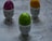 Three vibrantly colored eggs, one green, one purple, and one yellow, each resting in egg cups. The egg cups are decorated with different patterns, including red polka dots and yellow flowers. The background is a textured gray surface.