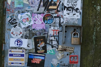 A metal surface covered with a variety of colorful stickers and graffiti. Some stickers display text, logos, or graphic designs, including anti-Nazi messages, music-related themes, and personal expressions. A solid padlock is attached to the right side, providing a contrast to the vibrant collage of the stickers.