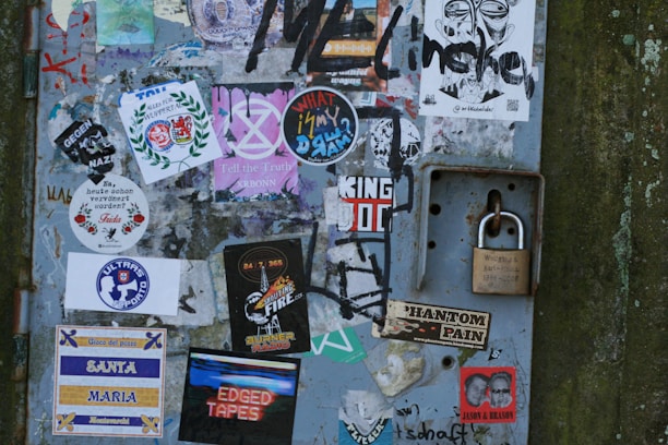 A metal surface covered with a variety of colorful stickers and graffiti. Some stickers display text, logos, or graphic designs, including anti-Nazi messages, music-related themes, and personal expressions. A solid padlock is attached to the right side, providing a contrast to the vibrant collage of the stickers.