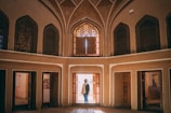 An interior view of a traditional Middle Eastern architectural structure with high arched ceilings and intricate geometric patterns. A person stands in the doorway, framed by colorful stained glass windows that cast vibrant patterns on the floor and walls. The architecture features symmetrical designs and warm tones, creating a sense of historical elegance.