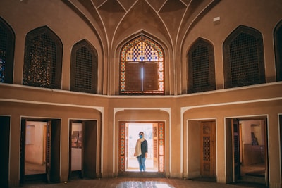 An interior view of a traditional Middle Eastern architectural structure with high arched ceilings and intricate geometric patterns. A person stands in the doorway, framed by colorful stained glass windows that cast vibrant patterns on the floor and walls. The architecture features symmetrical designs and warm tones, creating a sense of historical elegance.