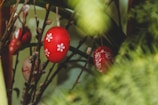 Colorful Easter eggs with floral designs hang from branches amidst a backdrop of green leaves. The eggs are primarily red with white flower patterns, creating a vibrant and festive look.